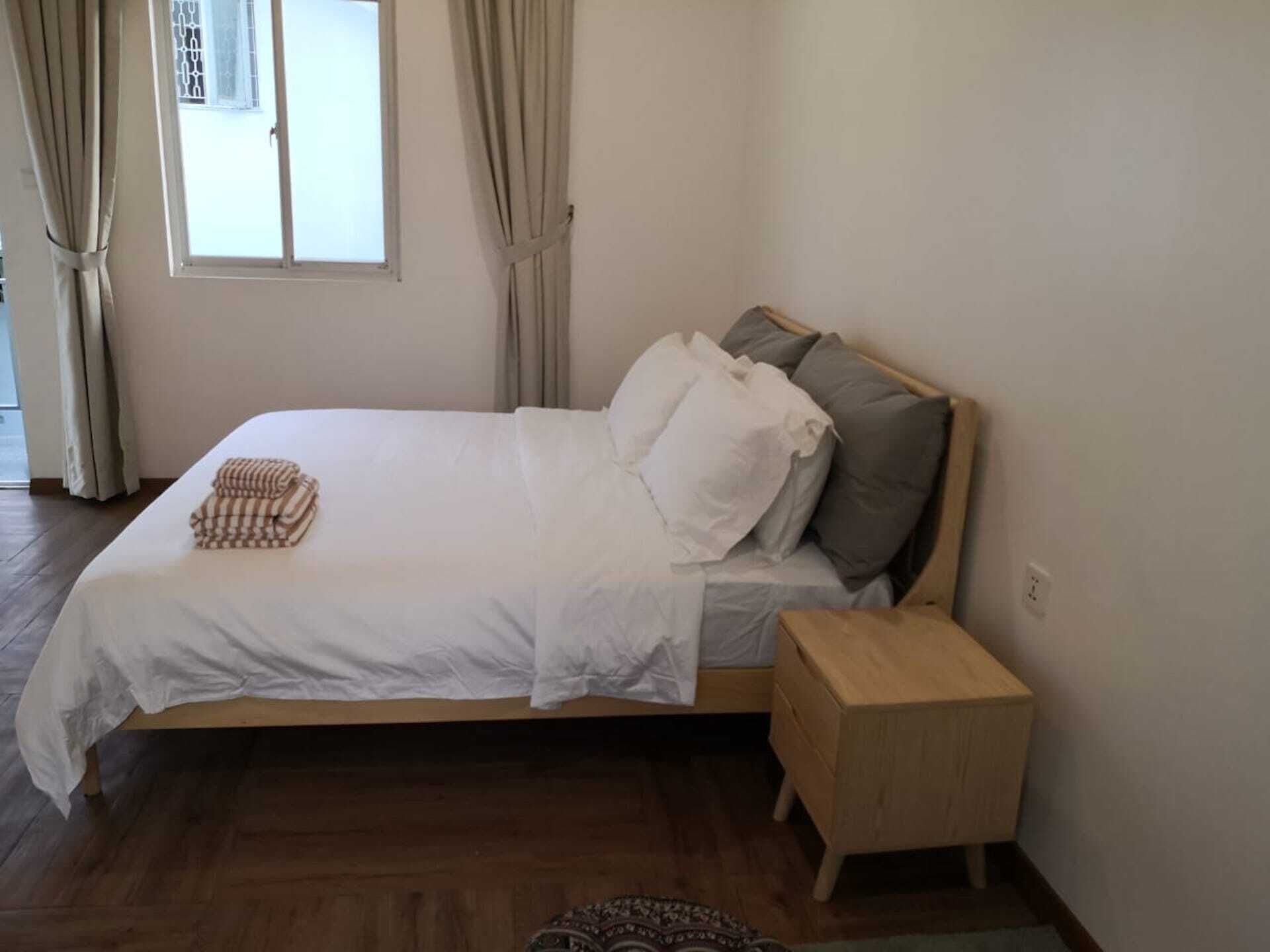 Een net opgemaakt bed met een houten hoofdbord, een nachtkastje en een raam met gordijnen.