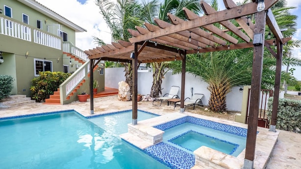 Une piscine dotée d’une pergola en bois et de bordures carrelées, entourée de palmiers et bordée d’un bâtiment à deux étages.