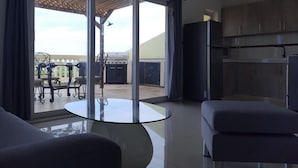 Un salon moderne avec une table en verre, deux canapés, un coin cuisine doté de placards et une porte-fenêtre coulissante donnant sur une terrasse extérieure avec un barbecue et des chaises.