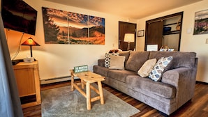 A cozy living room with a grey sofa, wooden coffee table, and a mountain landscape painting on the wall. - Cozy 1 Bedroom, Sleeps 3, Steps from Shuttle! Hot Tub! Loved by guests! (Crested Butte)