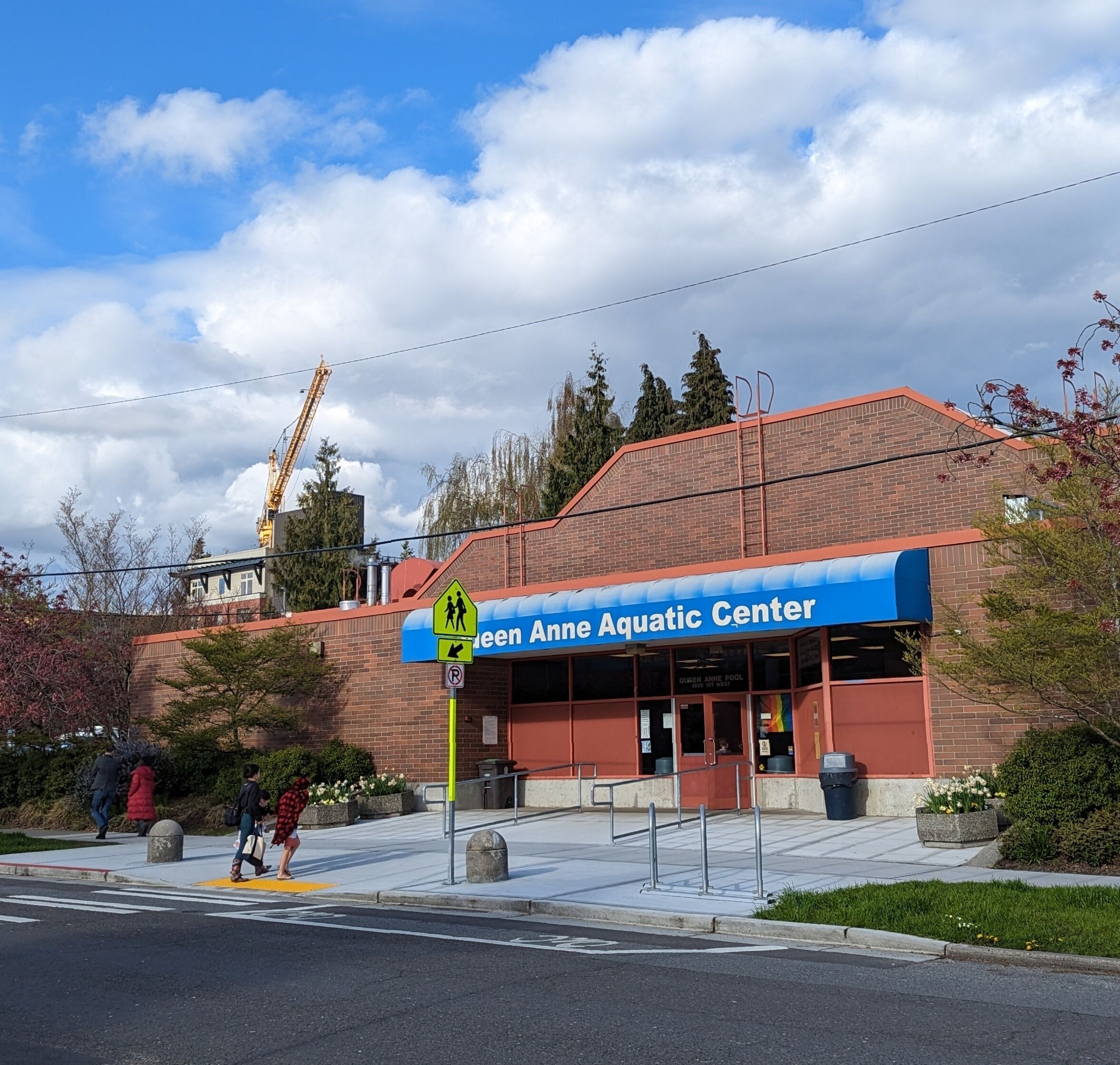The exterior of the Green Anne Aquatic Center with a blue awning, a pedestrian crossing, and a construction crane in the background.