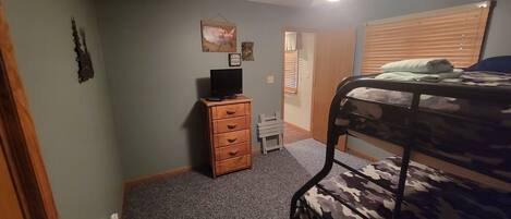 A bedroom with a bunk bed, a wooden dresser, a ceiling fan, and a door leading to another room.