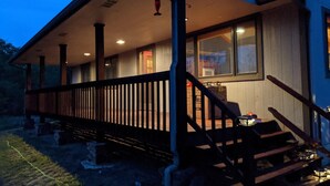 A covered porch with a wooden railing, a staircase leading to the entrance, and a view of the interior through the windows. - 2-bedroom vacation home sitting on your own private lake for fishing/relaxing. (Howard)