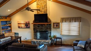 A cozy living room with a stone fireplace, a television mounted above it, and a dining area with a wooden table and chairs. - 2-bedroom vacation home sitting on your own private lake for fishing/relaxing. (Howard)