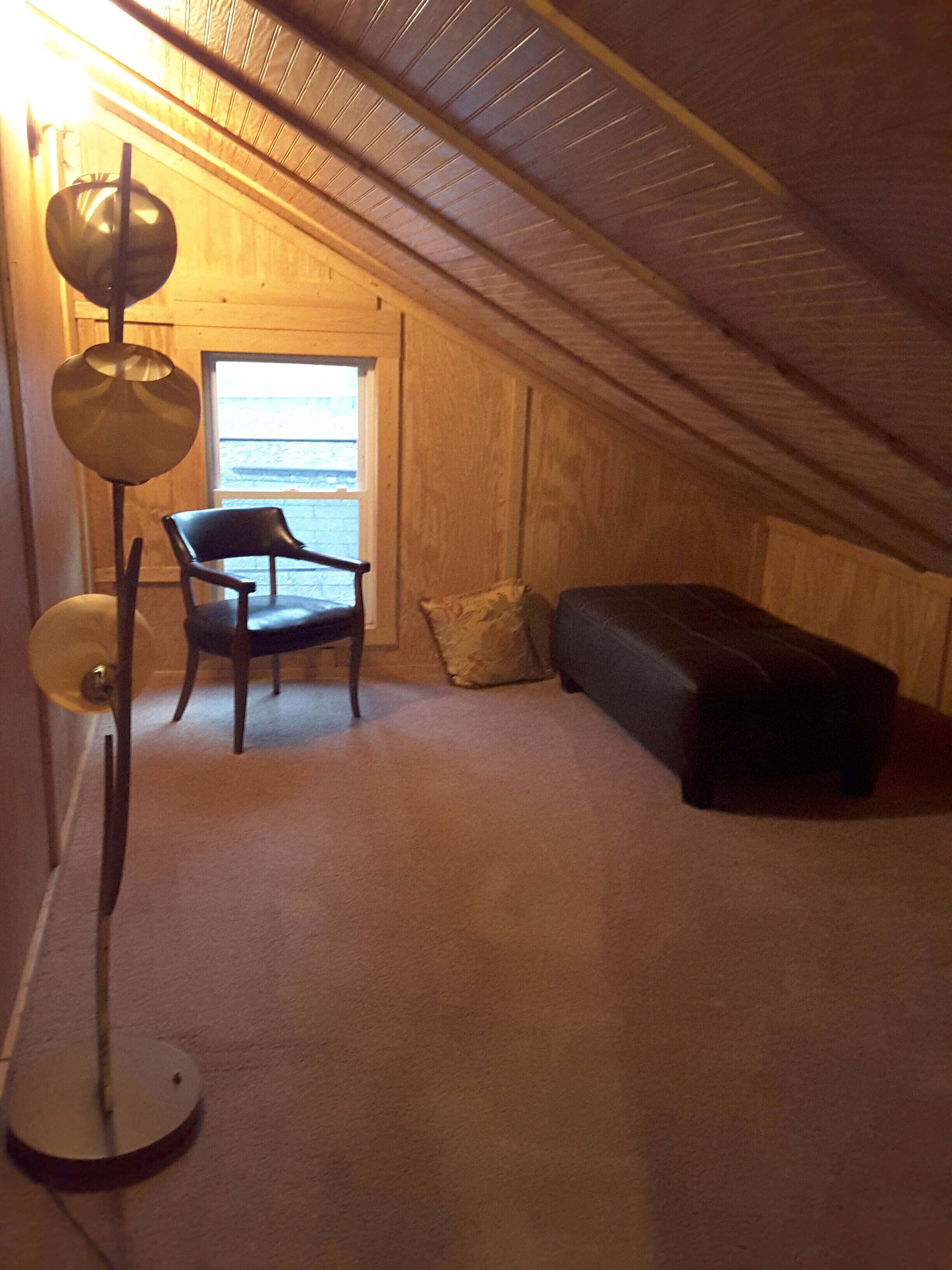 Ett litet vindsrum med ett sluttande tak, ett enda fönster, en stol, en soffa och en golvlampa.