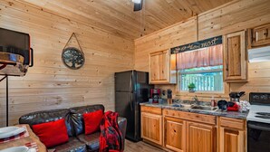 A cozy cabin-style kitchen with wooden walls, a black refrigerator, wooden cabinets, a sink, and a dining area with a leather couch and red cushions. - Cuddling On The Creek Couples only romantic " TINY CABIN" retreat!!!! (Cosby)