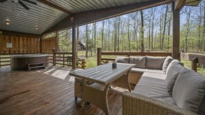 Um deck coberto ao ar livre com mesa de madeira e sofá de vime, uma banheira de hidromassagem e vista para uma floresta.