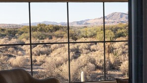 A room with a large window offering a view of a desert landscape. There is a sofa and a glass coffee table with a book and a basket on it.