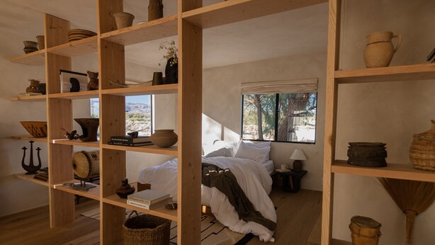 A room with wooden shelves displaying pottery and books, a bed with white linens, and a window with a view of trees.