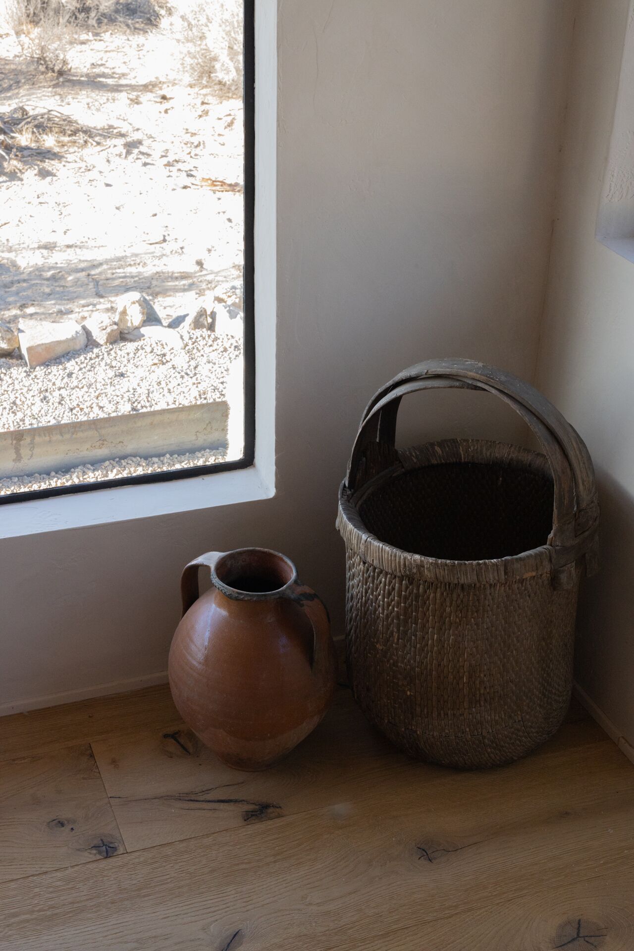 Una habitación rústica con suelo de madera, una maceta de barro y una cesta tejida cerca de una ventana.