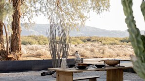 An outdoor seating area with wooden benches and tables, surrounded by gravel and desert vegetation.