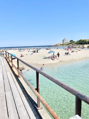 Eine Holzsteg führt zu einem Strand mit klarem Wasser, wo Menschen sonnenbaden, und im Hintergrund erhebt sich ein großes Gebäude.