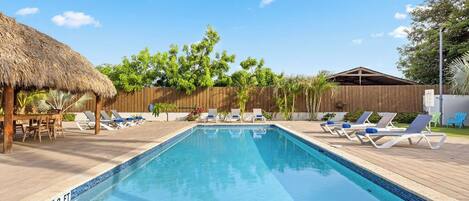 A rectangular swimming pool with a wooden deck, lounge chairs, and a thatched-roof structure.