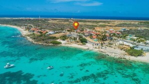 An aerial view of a coastal area with clear turquoise waters, a sandy beach, and a cluster of buildings with red roofs.