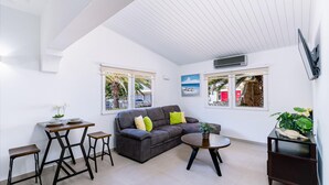A modern living room with a sofa, coffee table, and two stools. There are windows with views of palm trees and a beach.