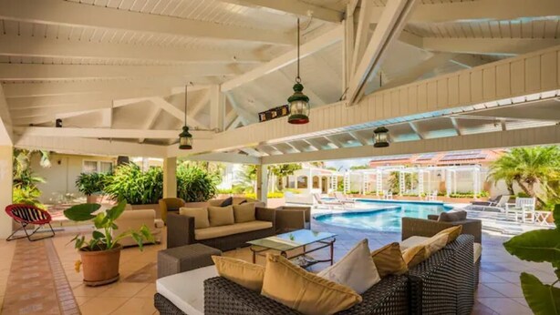 Un área de piscina interior con mobiliario de mimbre, plantas en macetas y vista a la piscina exterior.