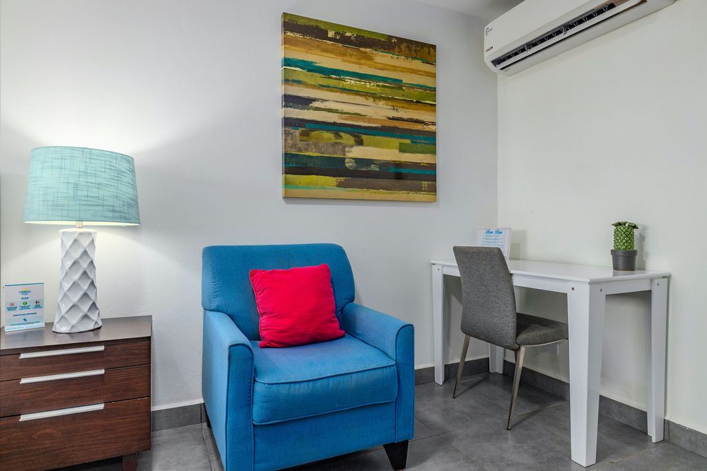 Ein Zimmer mit blauem Sessel, weißem Schreibtisch, Lampe und einem farbenfrohen, abstrakten Gemälde an der Wand.