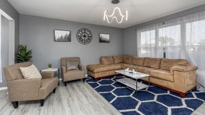 A living room with a sectional sofa, two armchairs, a coffee table, and a patterned rug. There are framed pictures on the wall and a ceiling light fixture.