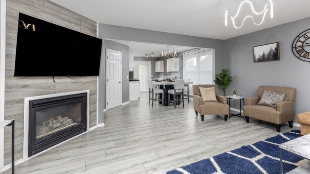 A modern living room with a fireplace, a flat-screen TV mounted on a wooden wall, a dining area with a table and chairs, and a kitchen in the background.