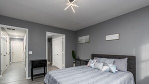 A bedroom with a bed, bedside table, and a nightstand. There is a door leading to another room and a hallway.