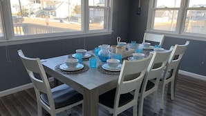 A dining table set with plates, bowls, and glasses, surrounded by white chairs, with a view of a street and buildings through large windows.