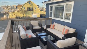 A covered outdoor seating area with wicker furniture and cushions, a glass-top table, and a view of a yellow house in the background.