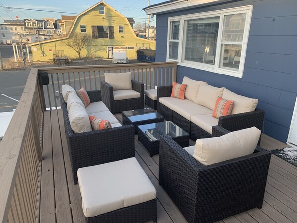 A covered outdoor seating area with wicker furniture and cushions, a glass-top table, and a view of a yellow house in the background.