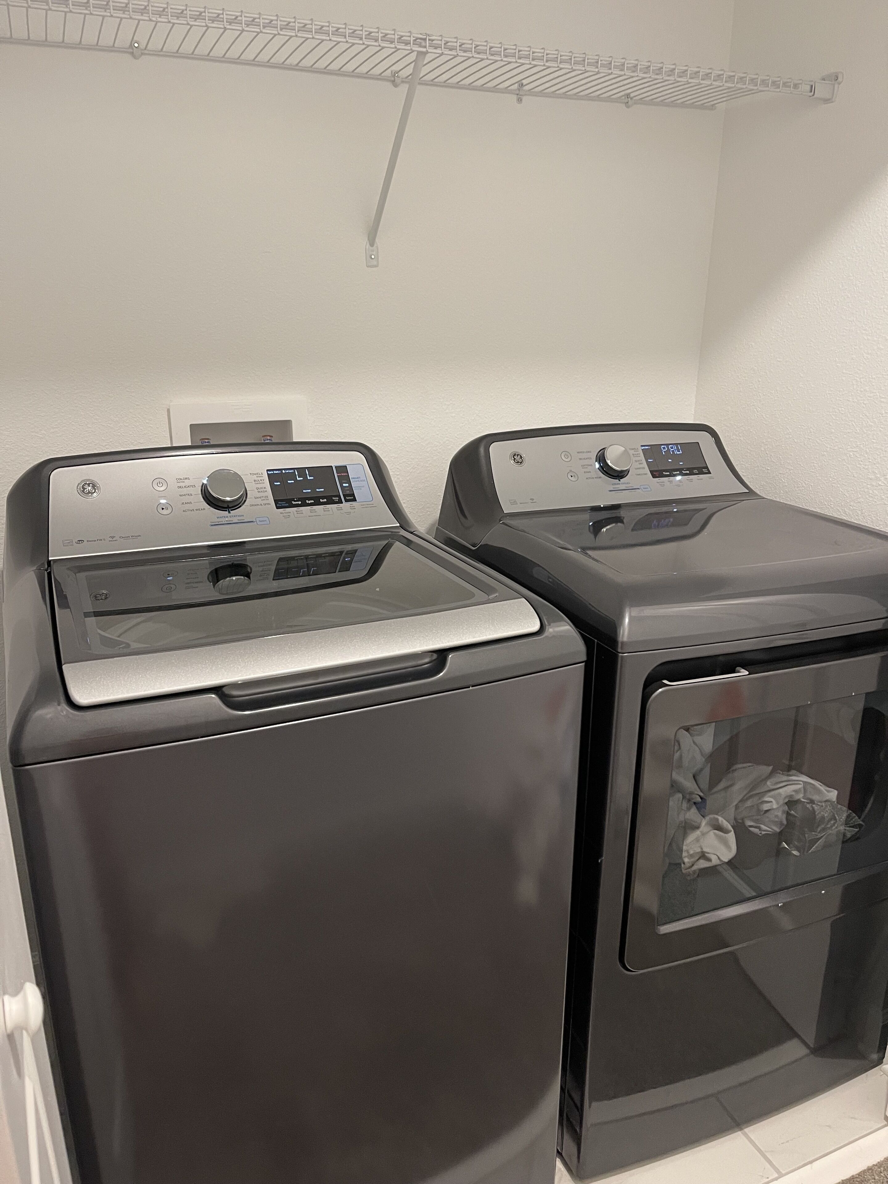 Two front-loading washing machines in a laundry room with a white wall and a metal shelf above.