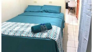 Une petite chambre avec un lit simple, une couvre-lit de couleur turquoise, un coussin à motifs et une serviette enroulée. On aperçoit une chaise et une petite table au fond de la pièce.