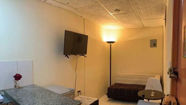 Une petite pièce compacte comprenant un coin cuisine, un lit, une table de chevet, une chaise et un téléviseur fixé au mur.