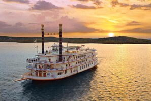 Un gran barco de recreo fluvial con varias cubiertas y una chimenea destacada, navegando sobre una masa de agua durante el atardecer.
