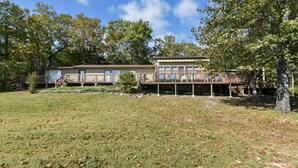 A two-story house with a deck, surrounded by trees and a grassy yard. - Hoot Owl @ Table Rock Lake - Amazing Lake Views!! - Game Room - Priced TO Rent!! (Kimberling City)