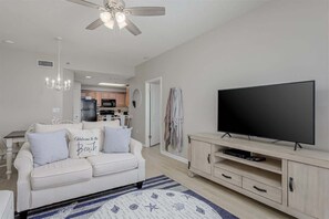A living room with a white sofa, a flat-screen TV, a ceiling fan, and a kitchen in the background.