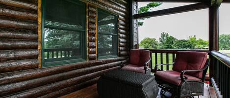 A log cabin with a wooden deck, two wicker chairs, and a wicker ottoman.