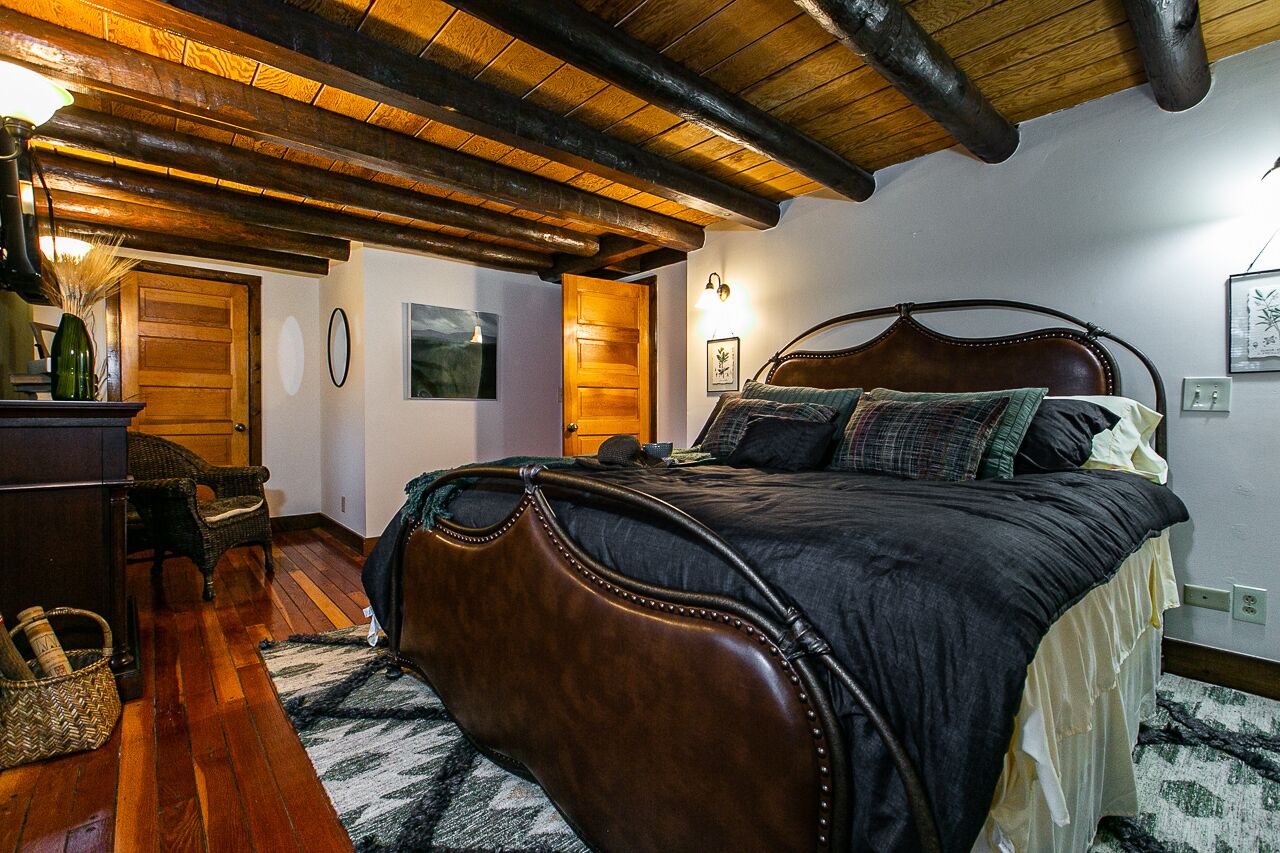 A bedroom with a wooden ceiling, a large bed with a headboard, a dresser, wicker chairs, and a rug.