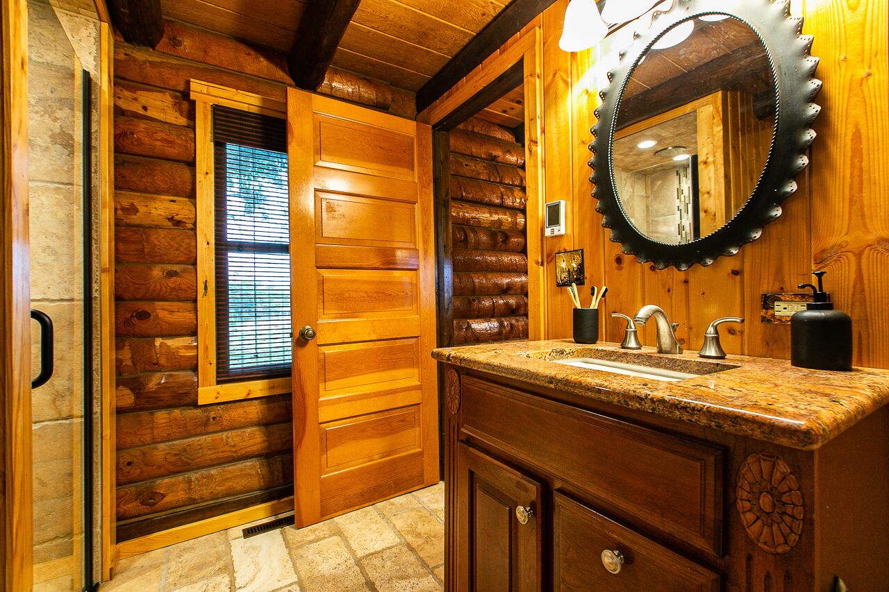 A wooden cabin bathroom with a granite countertop, a round mirror with a decorative frame, and a wooden door with a window.