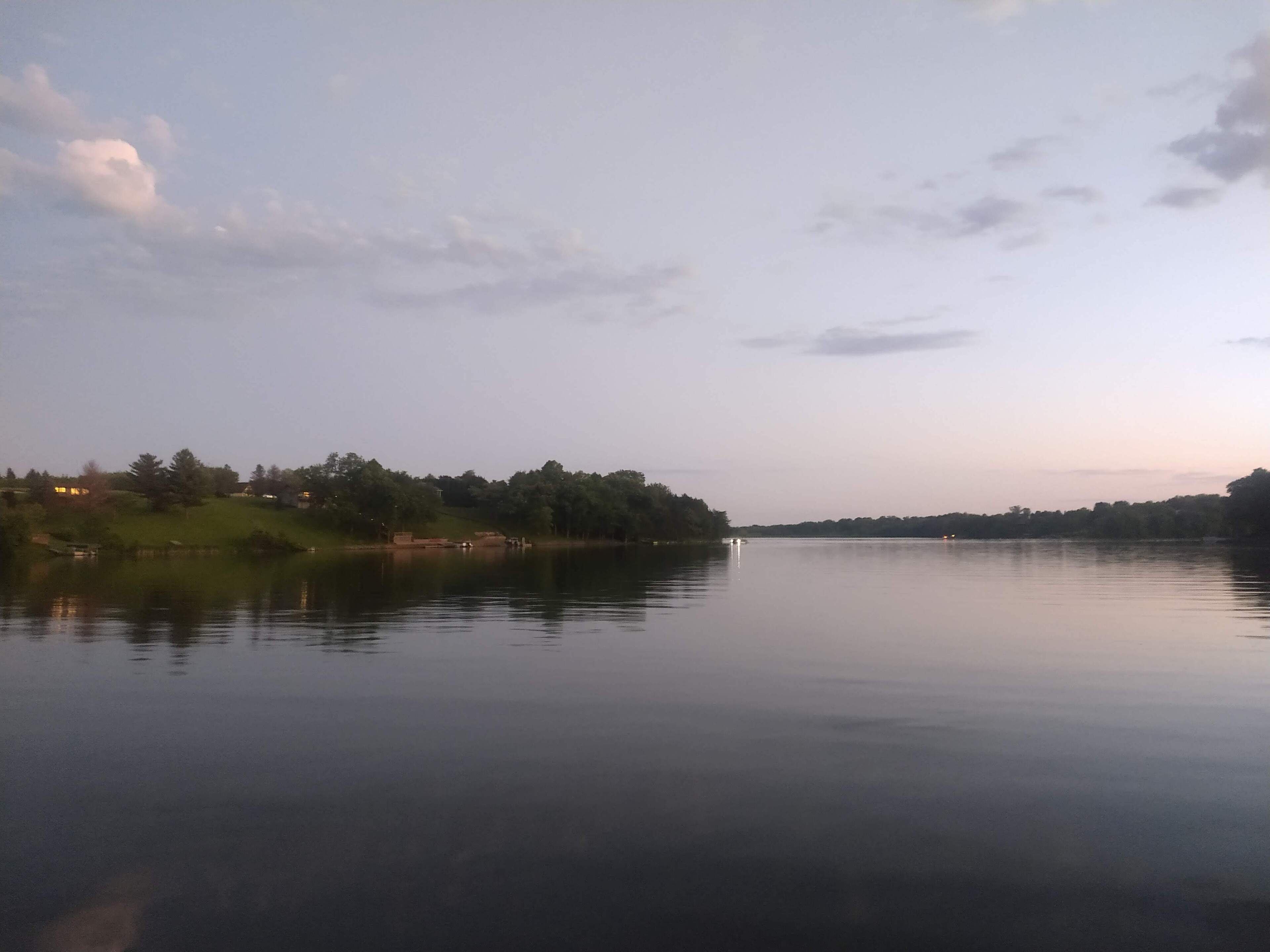 Un lac paisible, avec une rive lointaine, des arbres et quelques bâtiments.