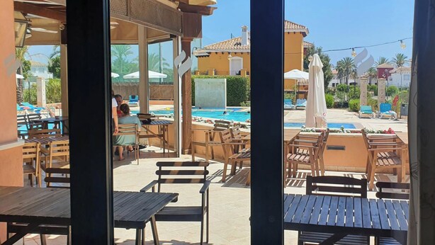 An outdoor patio area with wooden tables and chairs, a pool, and umbrellas. - Luxury Golf View 3Bed Penthouse, Sleeps 6, Balcony (Mar Menor Golf)
