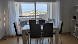 A dining area with a table and chairs, a bookshelf, and a view of the sea and buildings. - Luxury Golf View 3Bed Penthouse, Sleeps 6, Balcony (Mar Menor Golf)