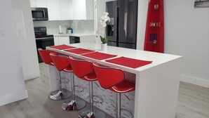 Uma cozinha moderna com armários brancos, uma geladeira de aço inoxidável, micro-ondas e um espaço para bar com banquetas vermelhas.