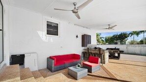 Um alpendre coberto com um sofá vermelho, duas cadeiras de vime, uma mesa pequena, ventiladores de teto e uma churrasqueira.