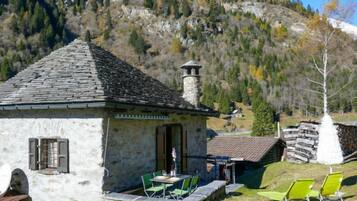 A stone building with a tiled roof, outdoor seating with green chairs, and a mountainous backdrop.