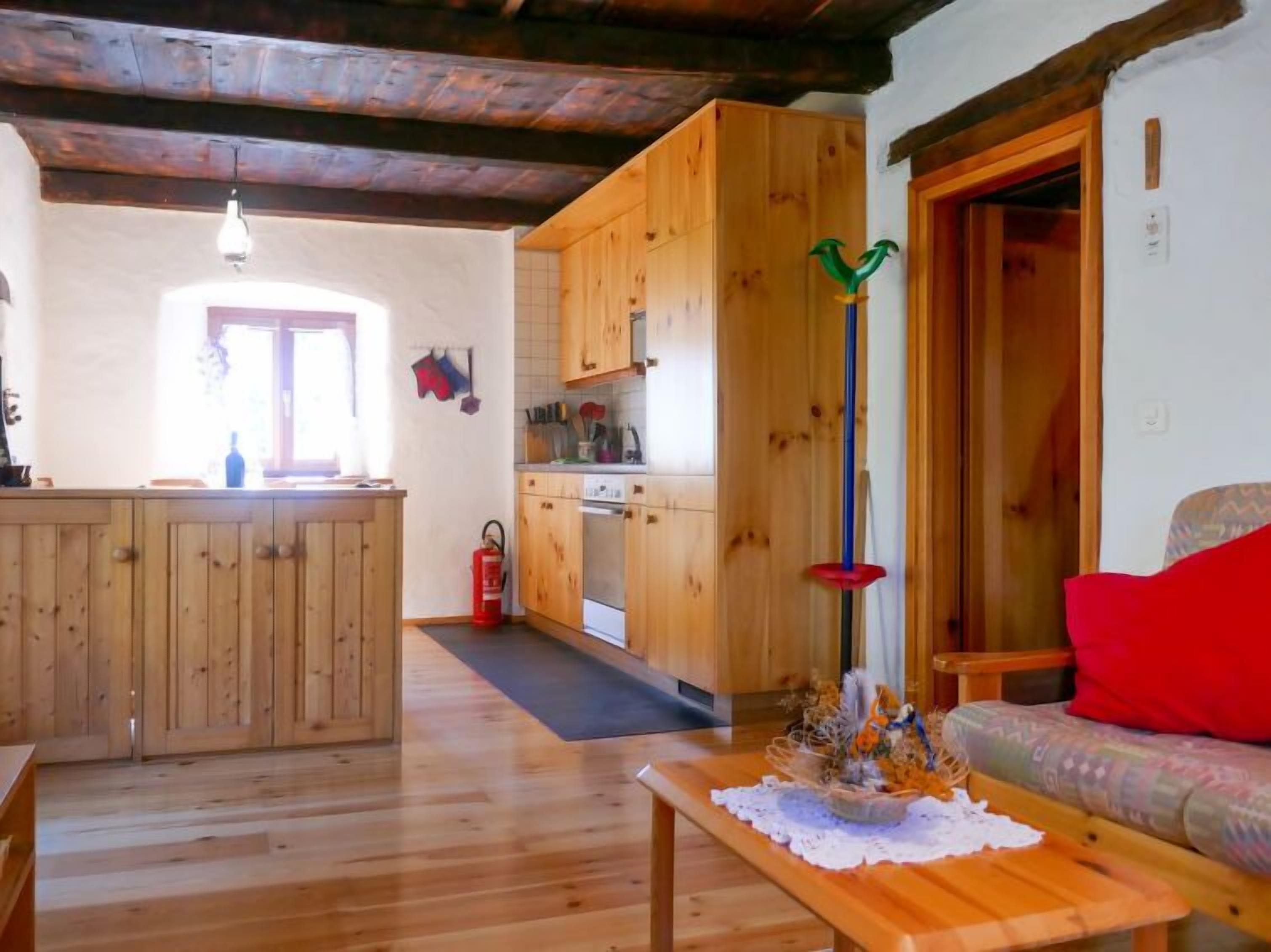 A cozy living room with a wooden bench, a dining table with a lace tablecloth, and a kitchen area with wooden cabinets and a stove.