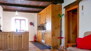 A cozy living room with a wooden bench, a dining table with a lace tablecloth, and a kitchen area with wooden cabinets and a stove. - Rustico Stübii by Interhome (Campo Blenio)