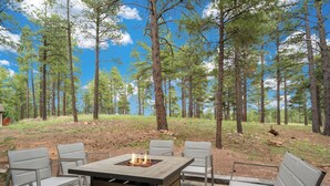An outdoor patio with a fire pit table, surrounded by chairs, set against a forest backdrop.