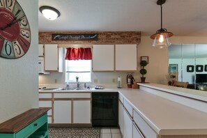 Une cuisine avec des meubles blancs, un lave-vaisselle noir, un lambrequin rouge à la fenêtre et une horloge au mur.