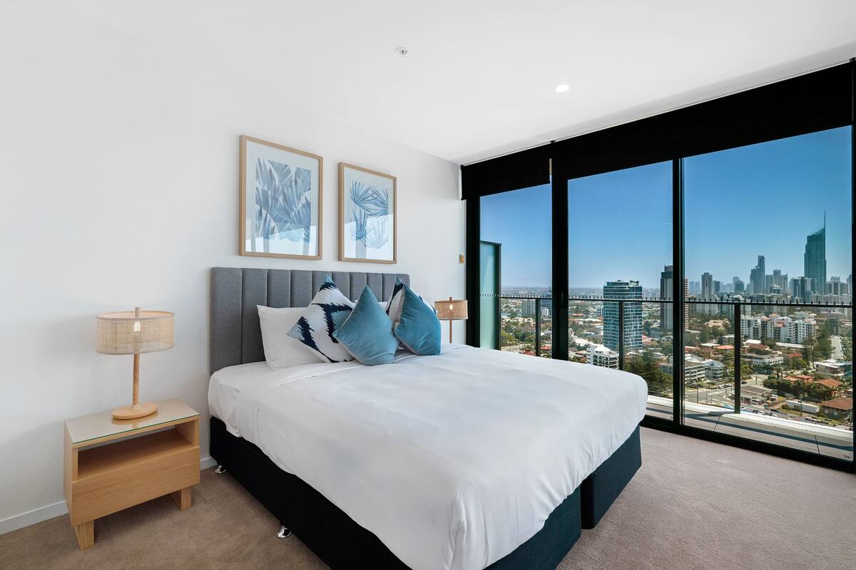 A modern bedroom with a large bed, a nightstand, and a city view through large windows.
