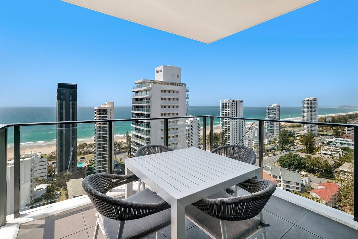 A balcony with a white table and chairs, overlooking a beach and cityscape.