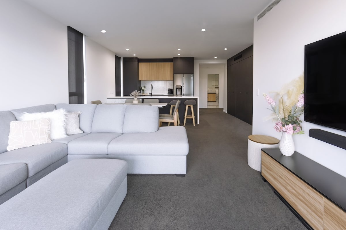 A modern living room with a grey sectional sofa, a flat-screen TV mounted on the wall, and a small round side table with a vase.