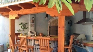 Een houten pergola buiten met een stenen tafel en vier houten stoelen. In de achtergrond is een stenen muur en een barbecue te zien.